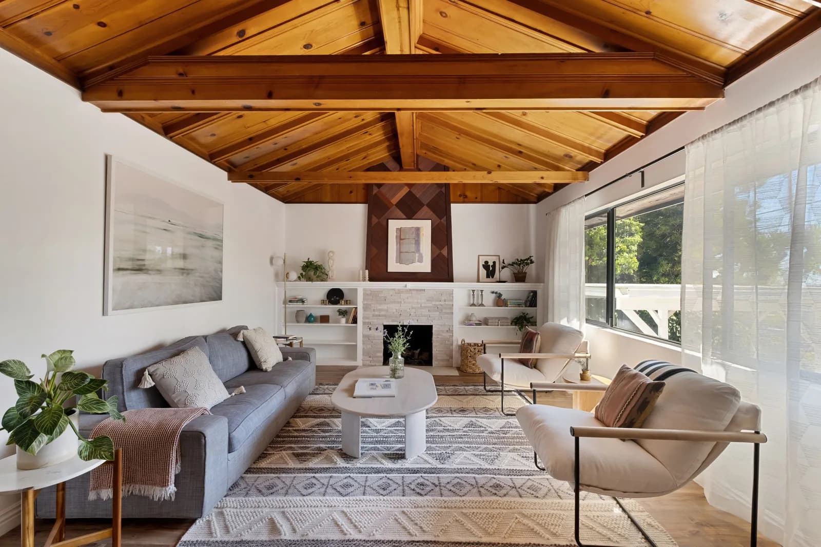 Staged living room with a vaulted wood-beam ceiling, stone fireplace, built-in shelves and a wide window
