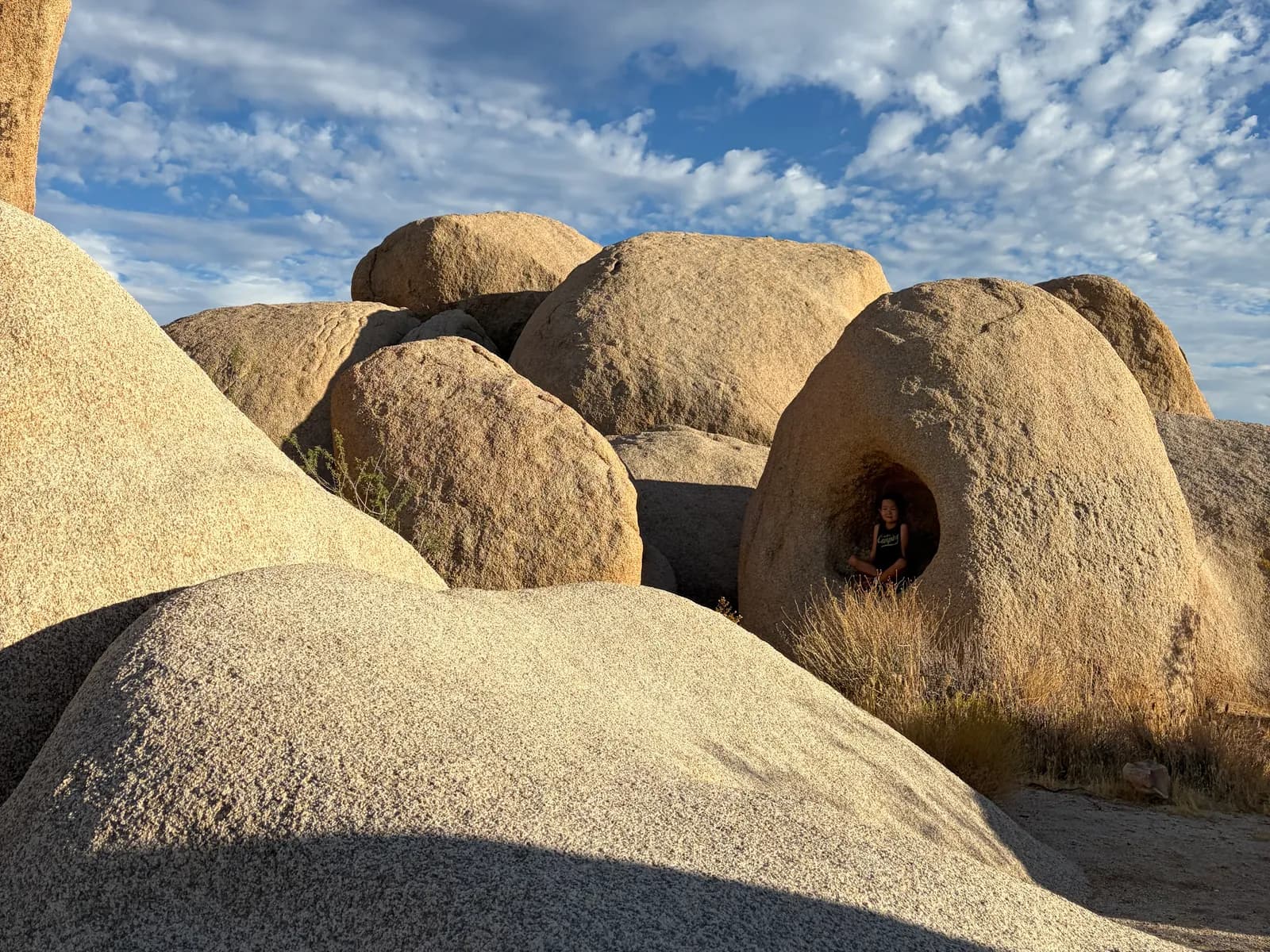 A kid sitting in a hollow of a giant desert boulder under a cloud-streaked sky