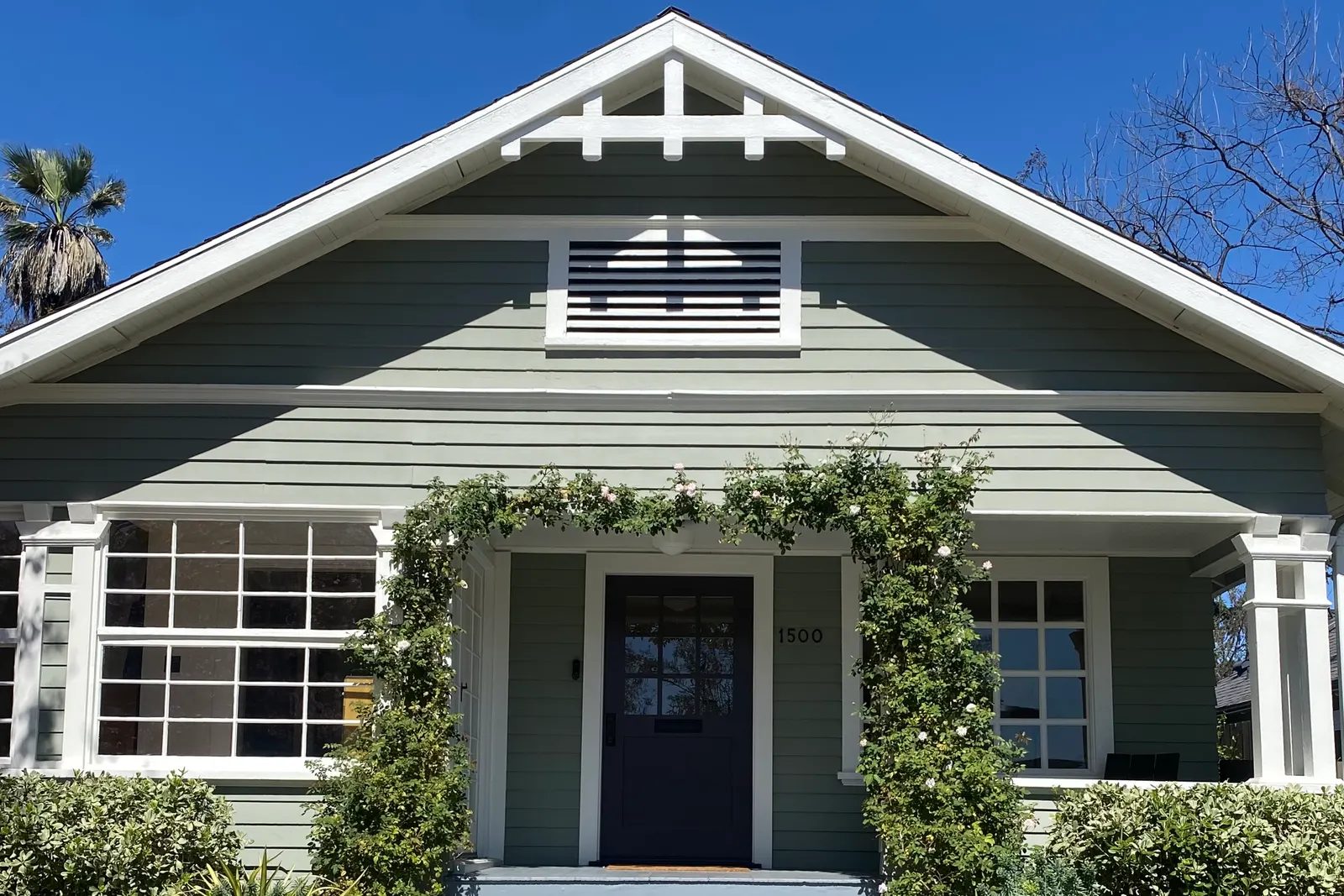 A sage-green Craftsman bungalow in South Pasadena with white trim, climbing roses over the porch and pale blue steps