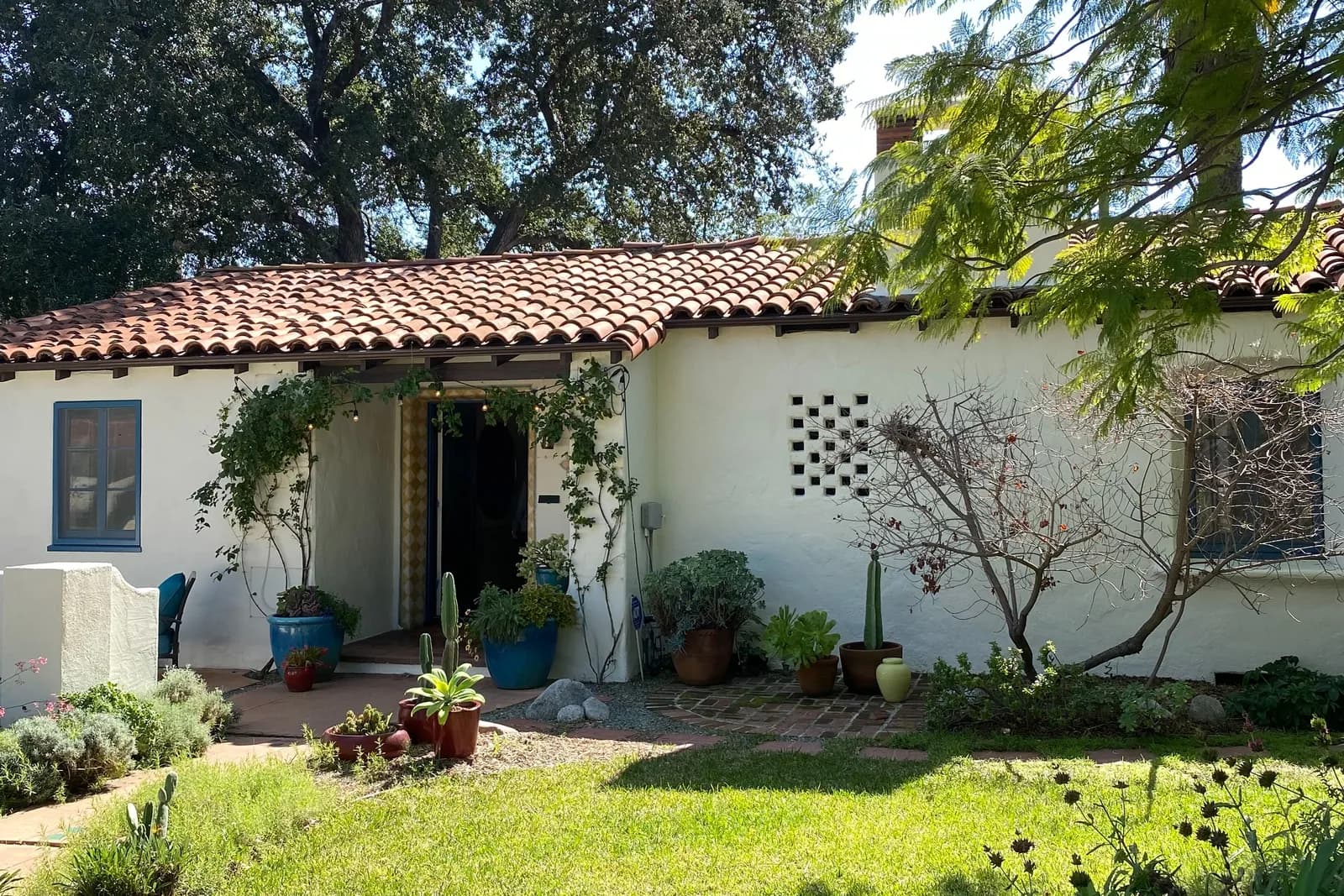 A white Spanish-style bungalow with a red tile roof, potted succulents and old oaks overhead
