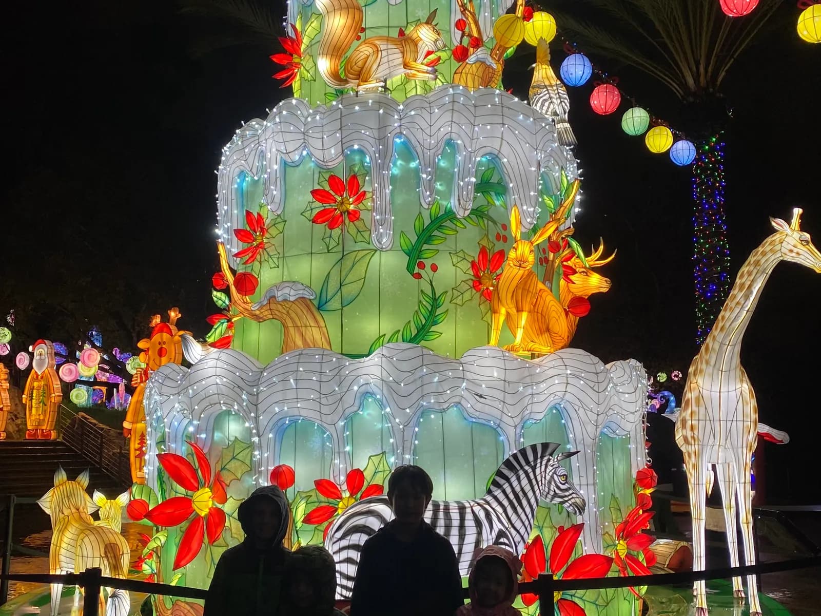 Kids silhouetted in front of a glowing lantern cake and giraffe at the L.A. Zoo holiday lights